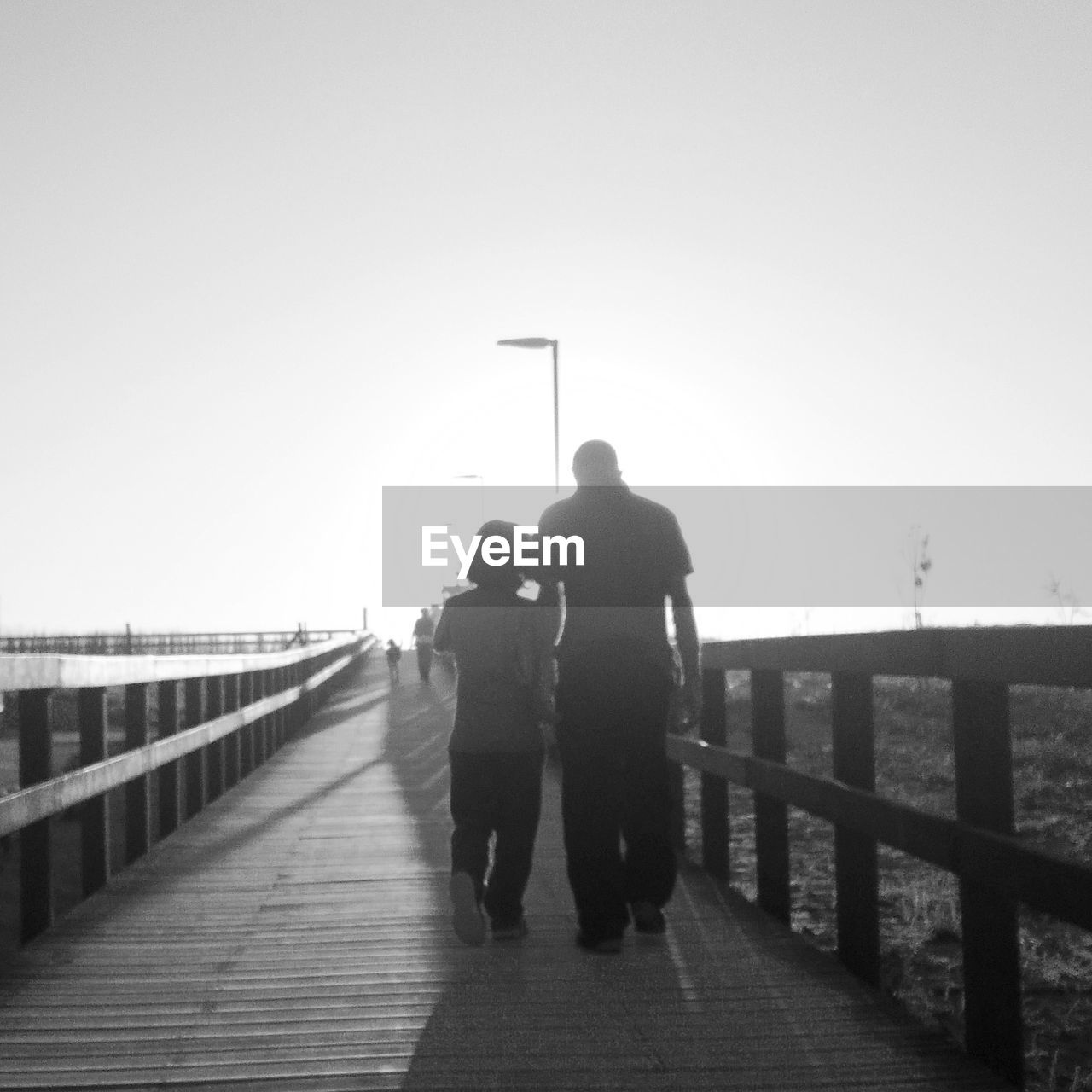 REAR VIEW OF MAN WALKING ON FOOTBRIDGE OVER RIVER