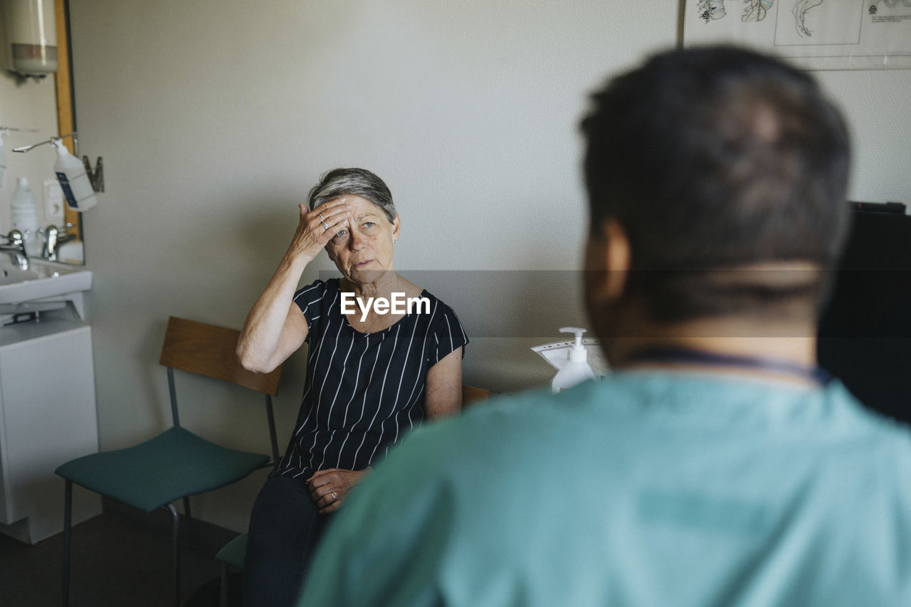 Senior woman touching forehead while discussing with doctor in clinic