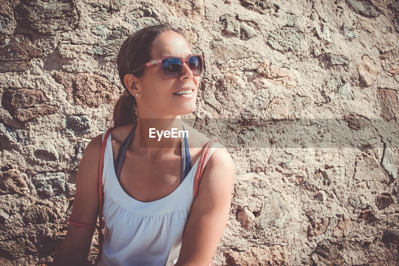 Smiling woman wearing sunglasses against wall during sunny day