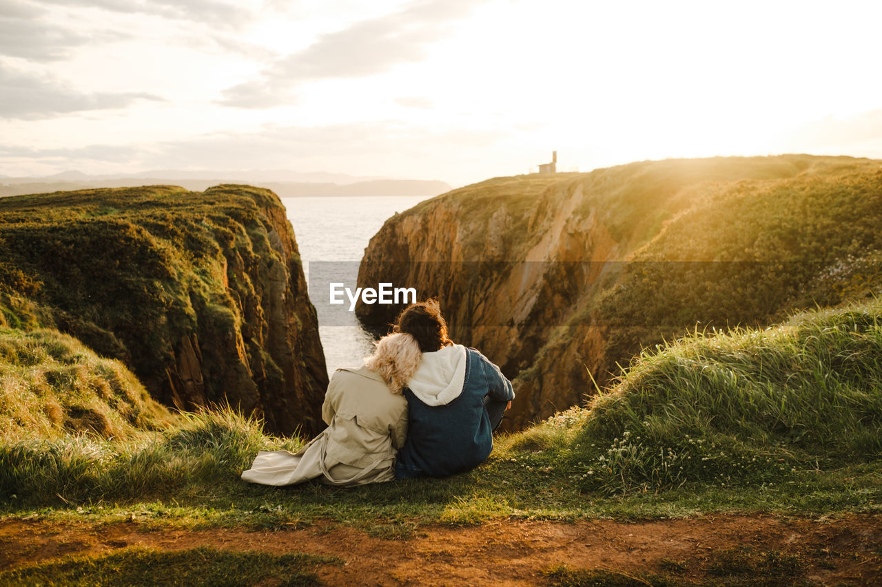 Back view of anonymous man and woman in outerwear leaning on each other and observing sea while sitting on cliff at sundown in aviles, spain