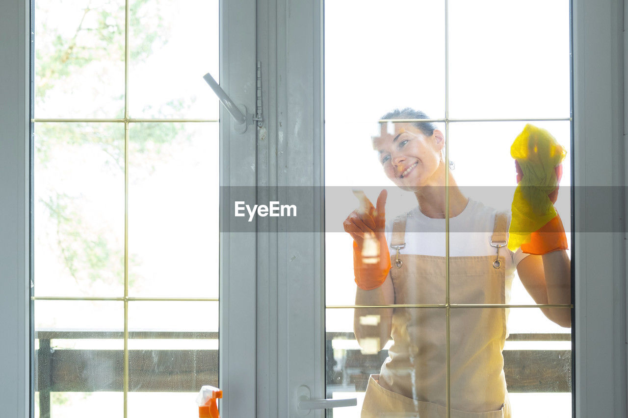 window, indoors, adult, one person, glass, standing, occupation, yellow, working, men, day, room, holding, interior design, business, person, protection, waist up, domestic room, transparent, communication, front view, mature adult, technology, business finance and industry, construction industry, looking, window covering, clothing, headwear, helmet, copy space, door, young adult, smiling, home interior, sunlight, women, back lit, office