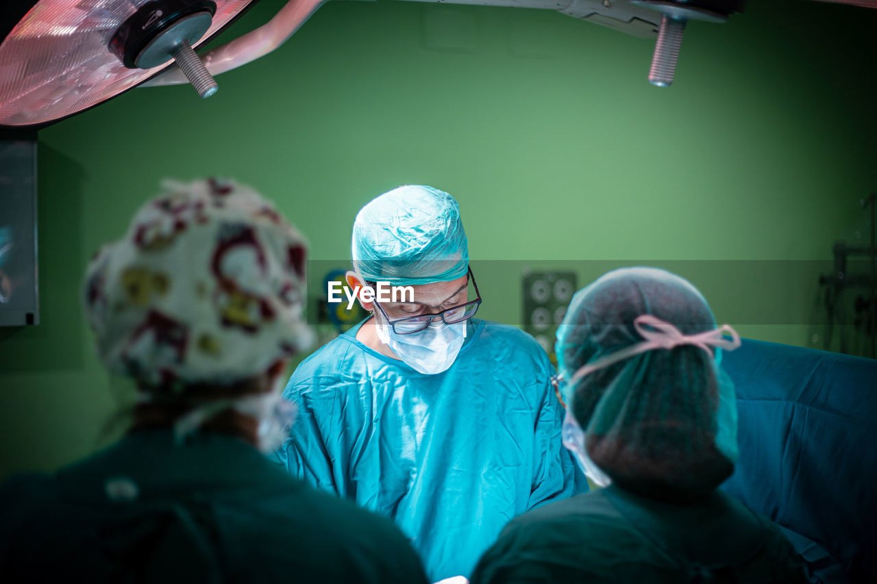 Male doctor in glasses and uniform performing surgery on patient with help of assistants in modern operating theater of hospital