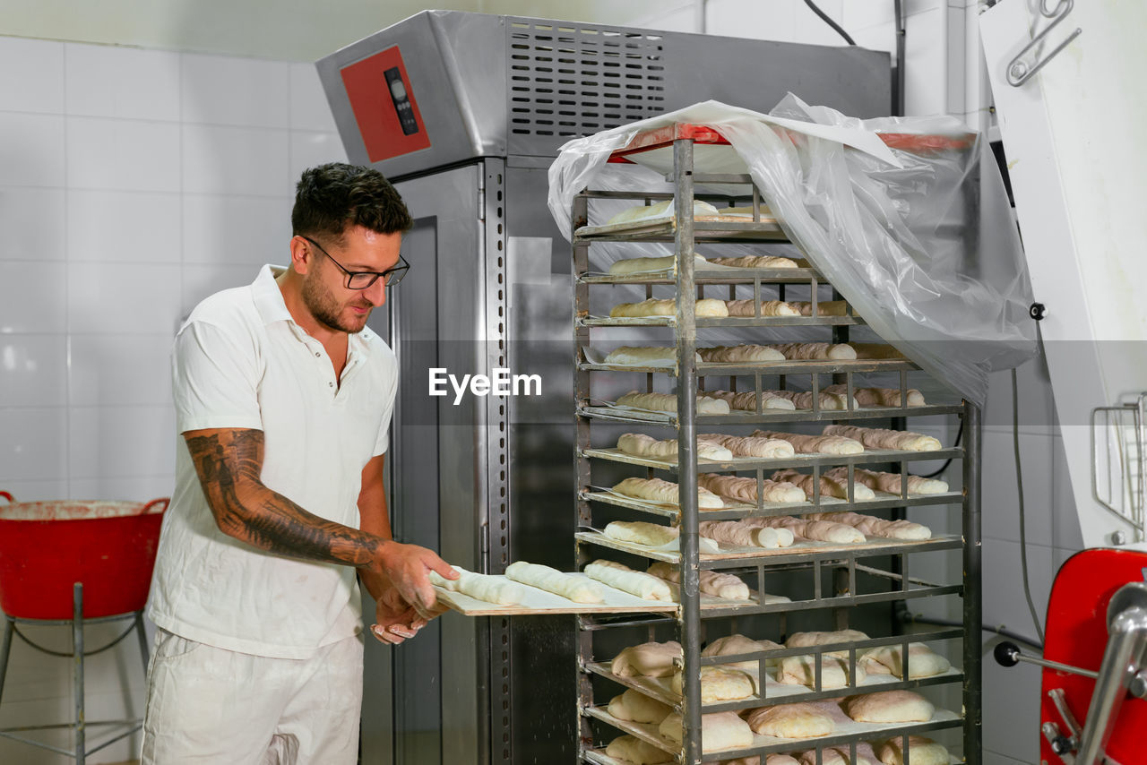 Young stylish tattooed male baker in casual clothes and eyeglasses putting loaves of raw dough on metal rack while baking bread in kitchen