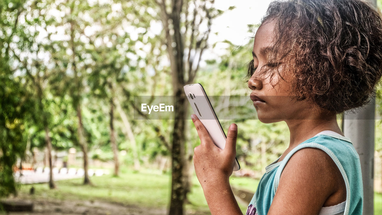 Side view of girl holding mobile phone against trees