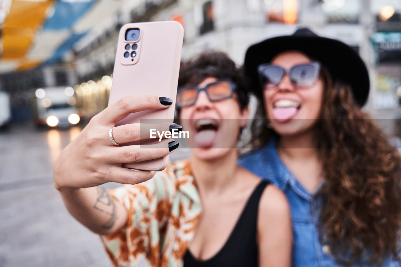 Soft focus of happy young girlfriends showing tongues and taking selfie via smartphone on weekend day on street on madrid, spain