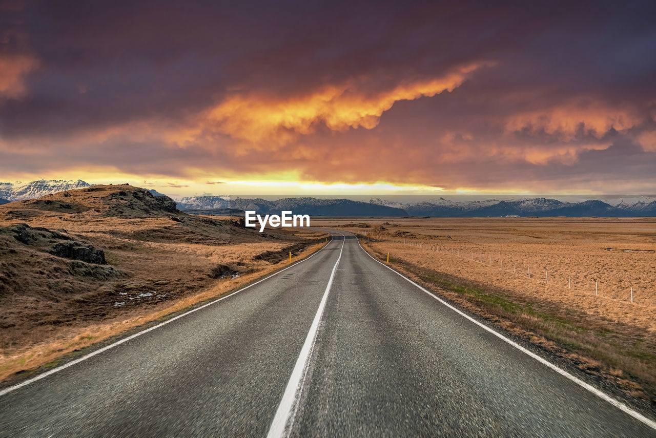 Empty vanishing street amidst volcanic landscape against dramatic sky at sunset