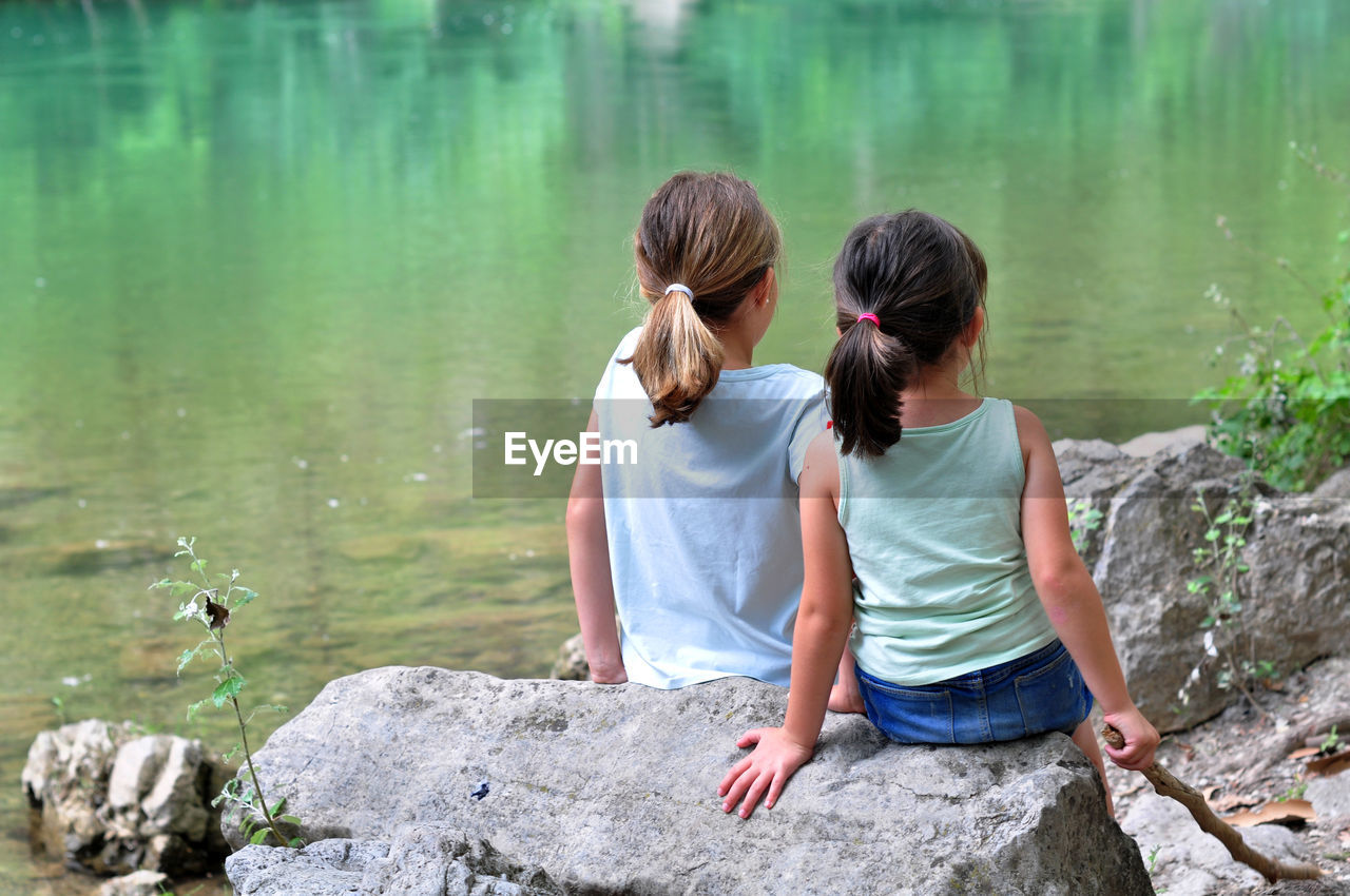 REAR VIEW OF WOMEN SITTING ON ROCK AGAINST LAKE