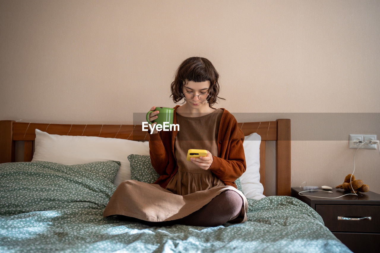 Pensive teenager sitting on bed with cup of tea, reading news in internet, hanging in social network