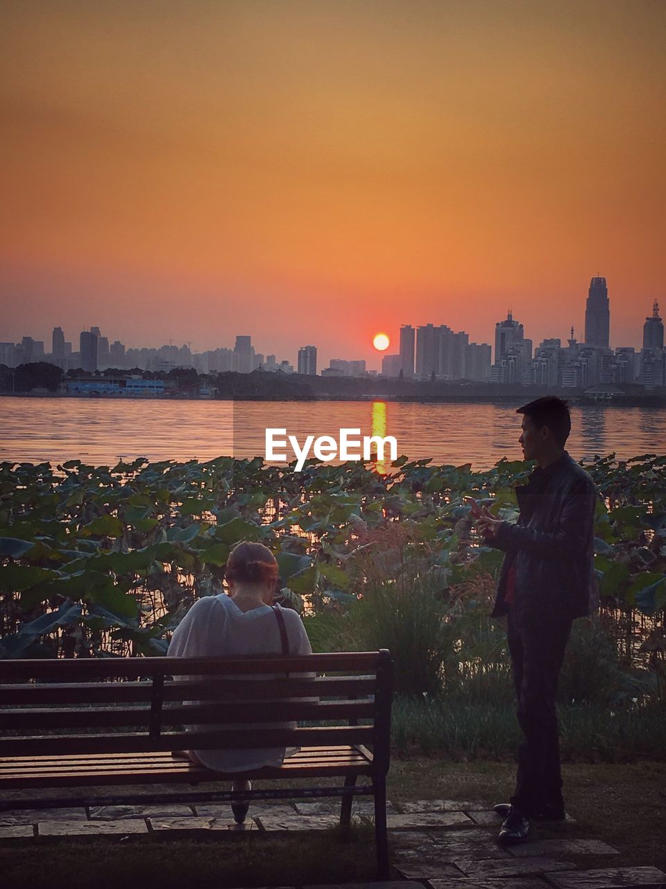 REAR VIEW OF MAN SITTING AT SEA AGAINST CITYSCAPE