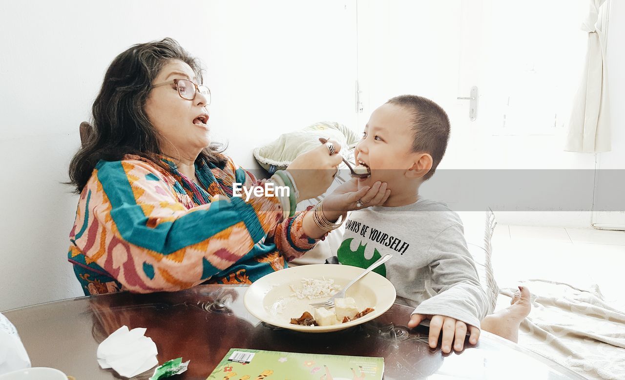 Mother feeding son while sitting by table at home