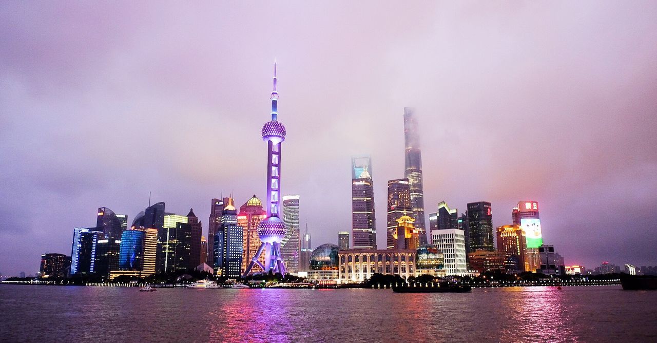 Illuminated skyscrapers by sea against cloudy sky at twilight