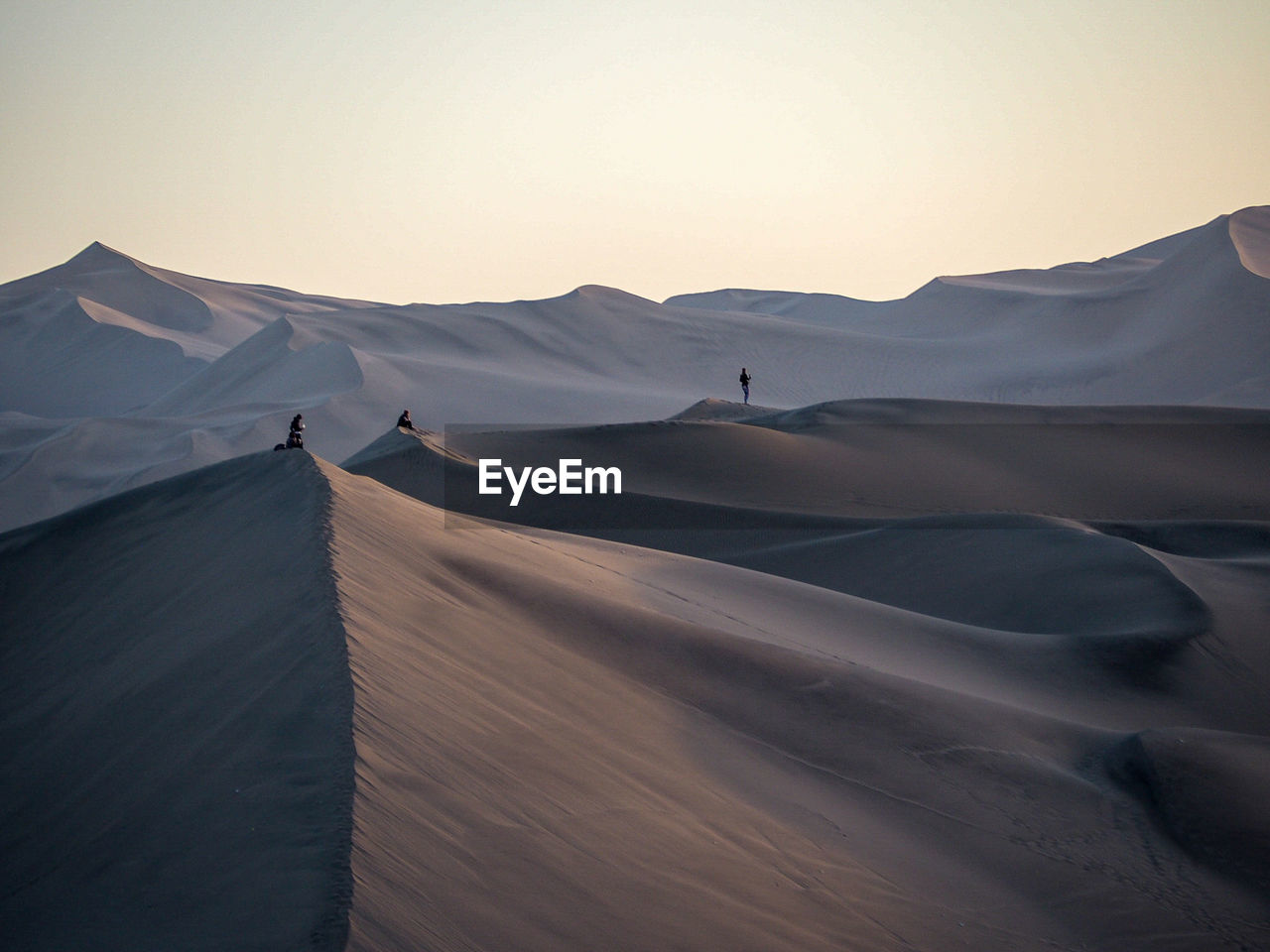 Mid distance view of people standing on sand dunes against clear sky during sunset