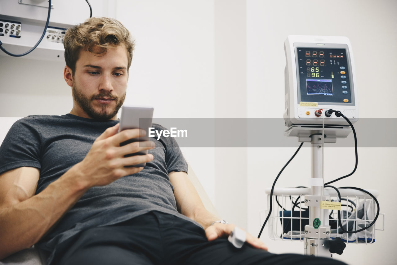 Young male patient using smart phone while reclining on bed during medical test in hospital