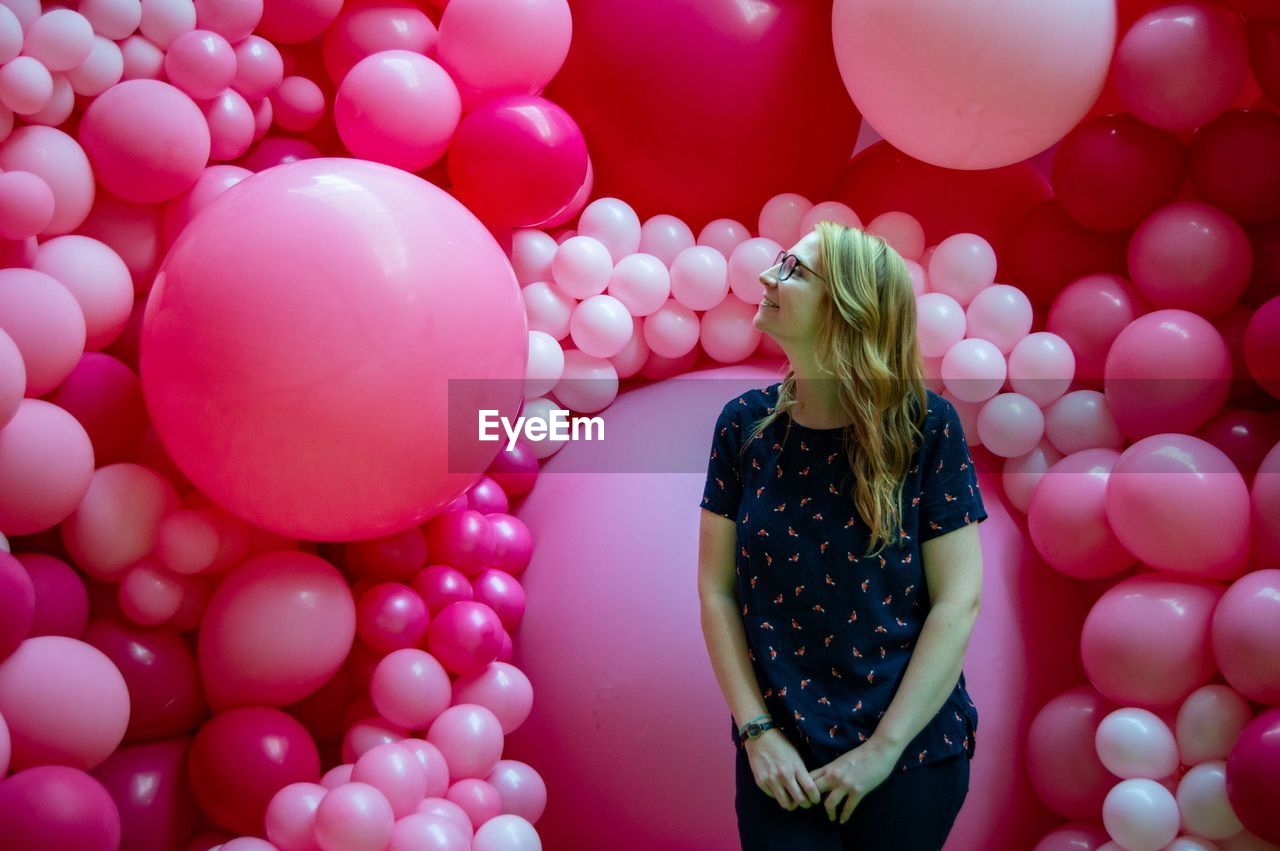 Smiling young woman looking away while standing against pink balloons