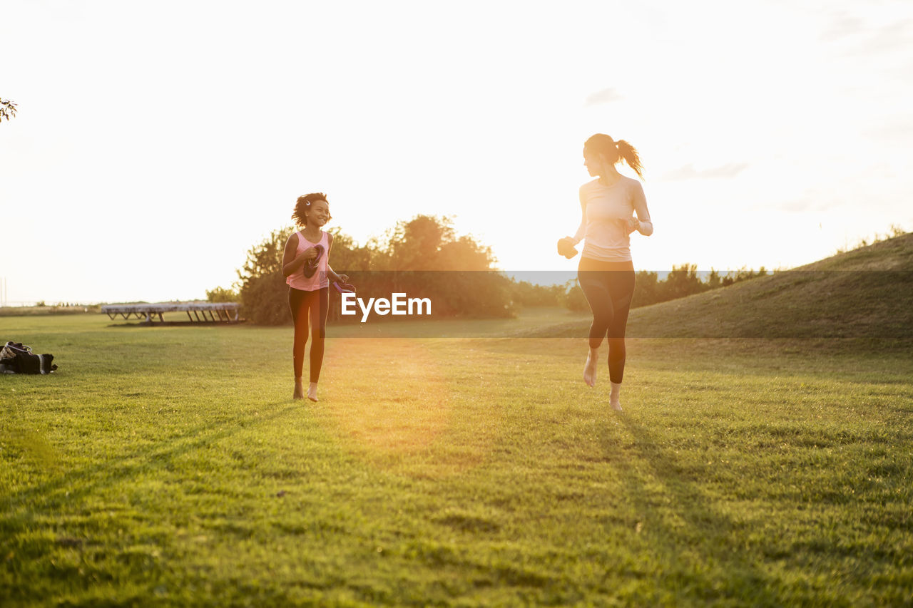 Happy girl jogging with mother on grass at park against sky during sunset