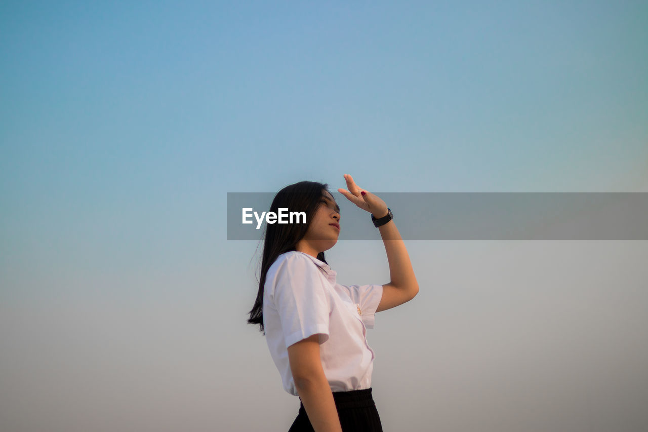 Low angle view of young woman shielding eyes standing against sky during sunset