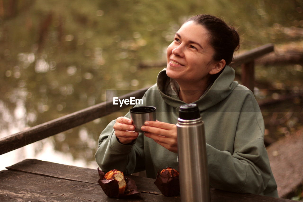 Young woman smiling drinking tea from thermos with muffin outdoors
