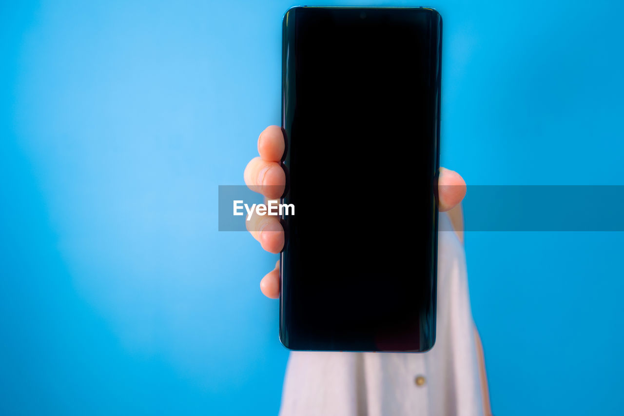 CLOSE-UP OF HAND HOLDING SMART PHONE AGAINST BLUE BACKGROUND