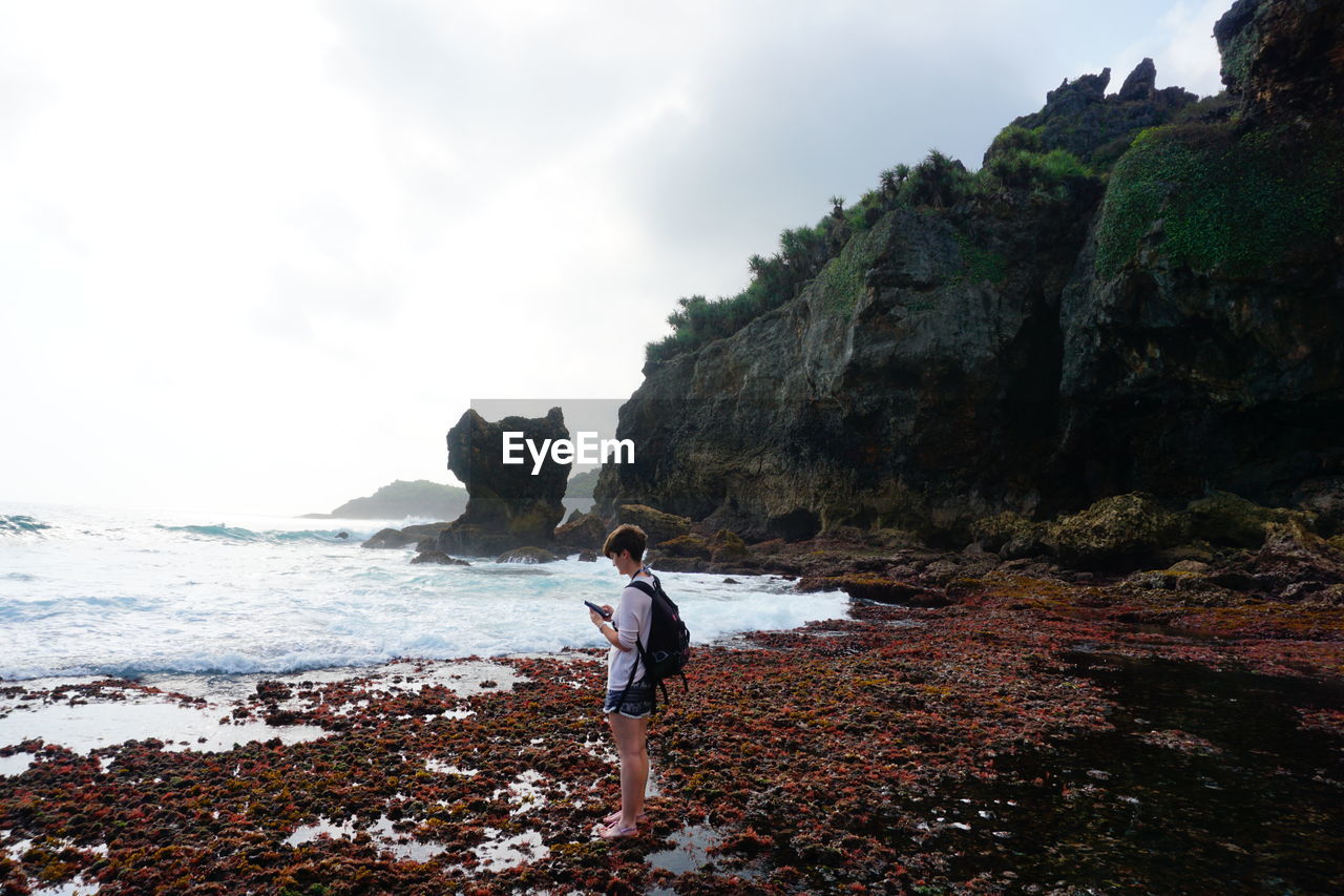 MAN STANDING ON ROCK LOOKING AT SEA AGAINST SKY