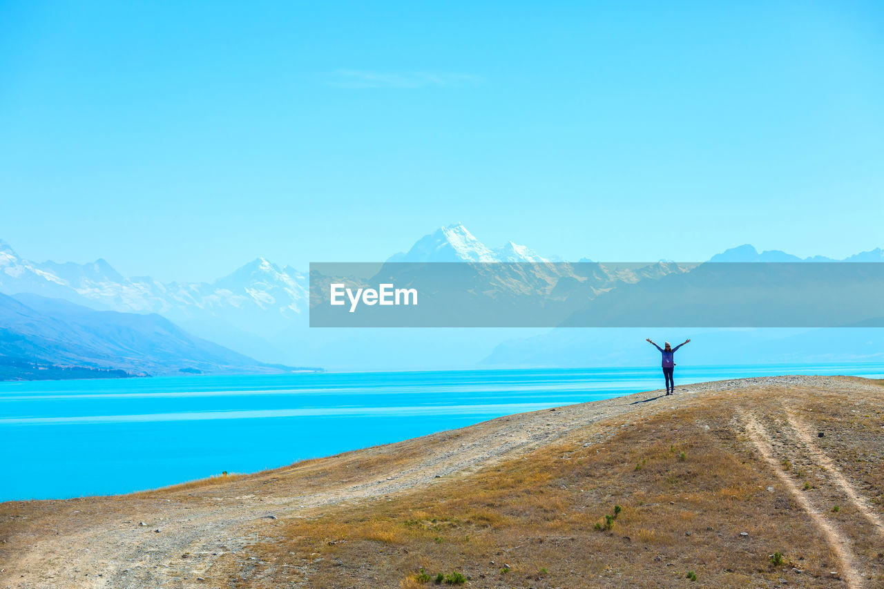 Woman with arms outstretched standing at lakeshore against mountains