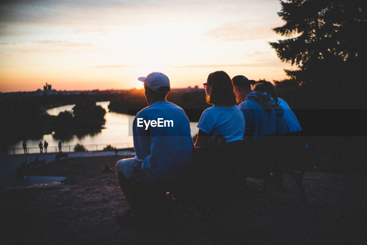 REAR VIEW OF PEOPLE SITTING ON SHORE AGAINST SUNSET SKY