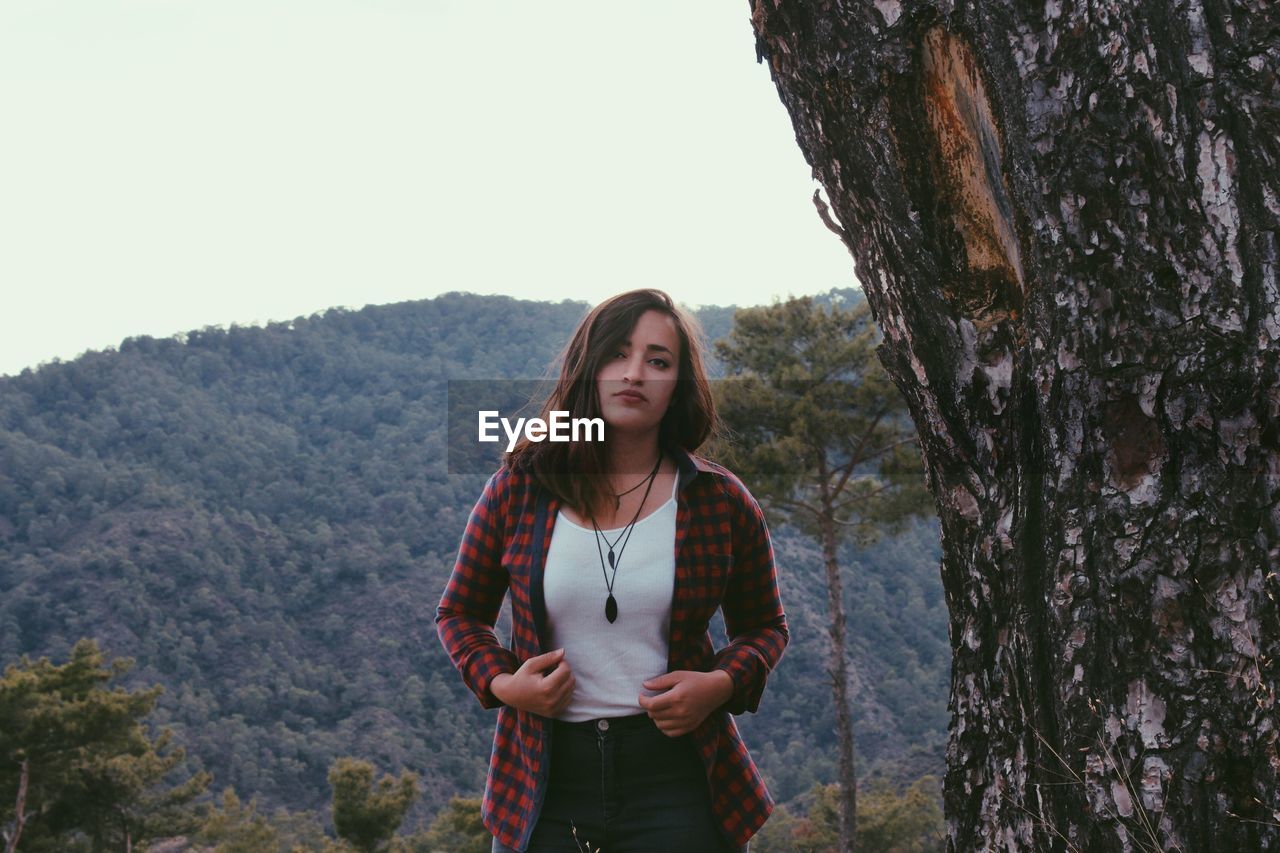 Portrait of young woman standing by tree against sky