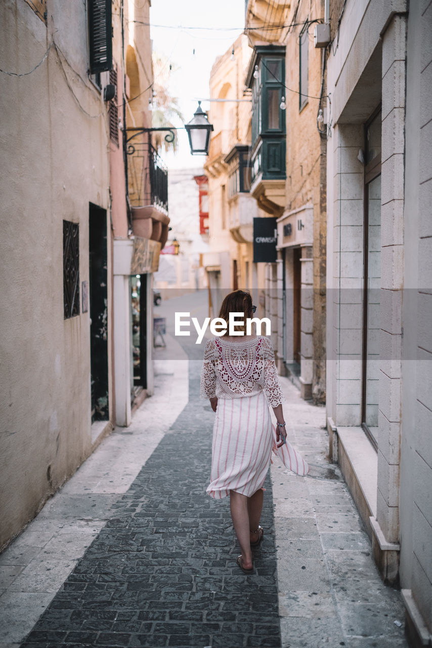 FULL LENGTH REAR VIEW OF WOMAN WALKING ON FOOTPATH AMIDST BUILDINGS