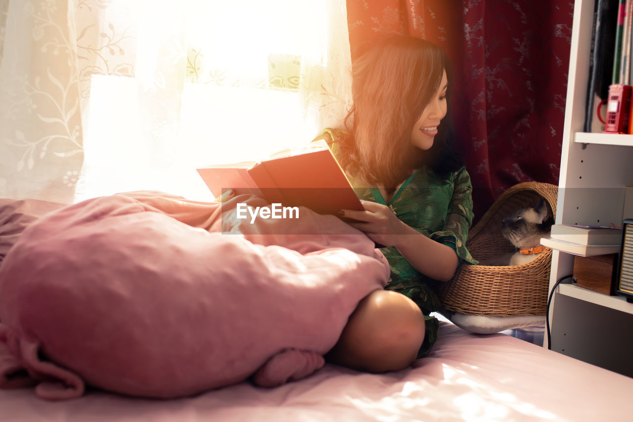 Woman reading book while sitting on bed by cat in basket at home