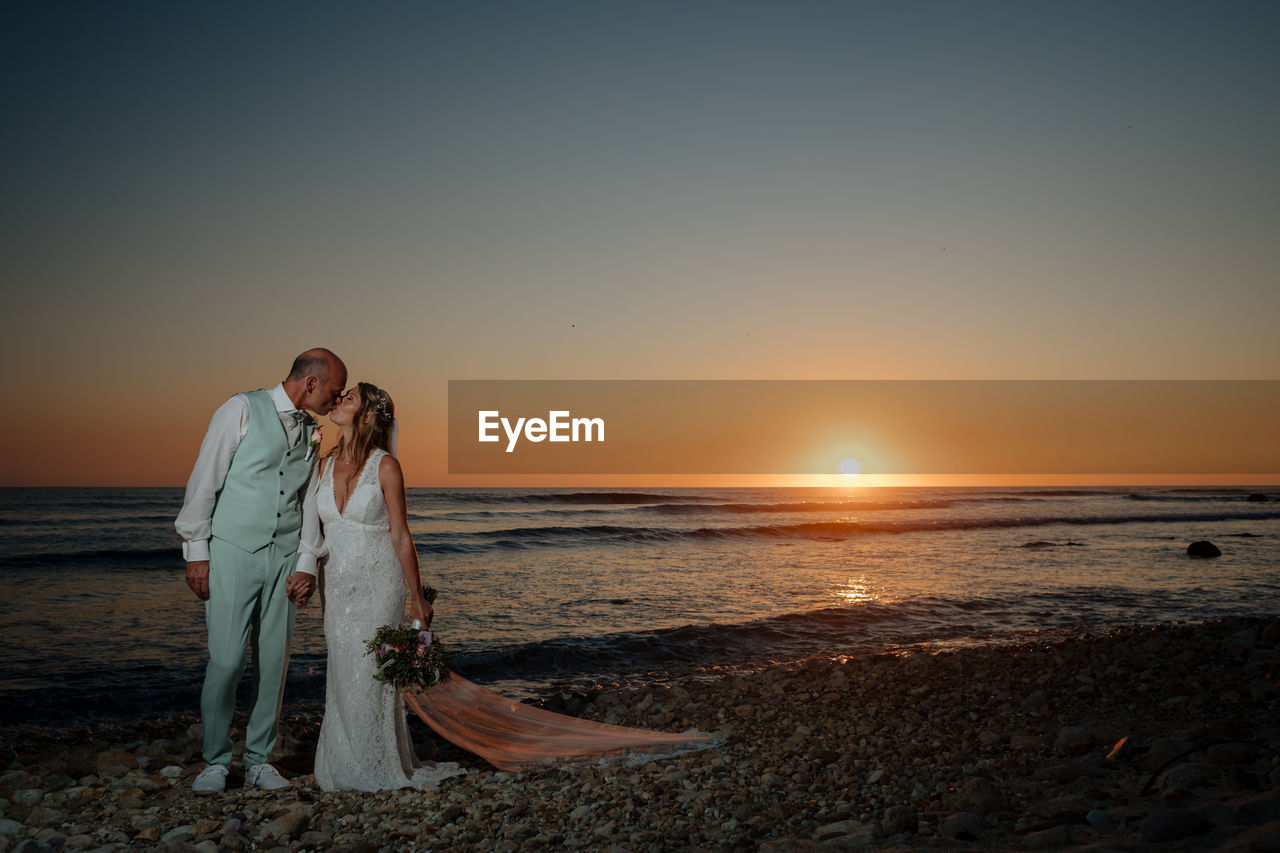 Bride and groom at beach against sky during sunset