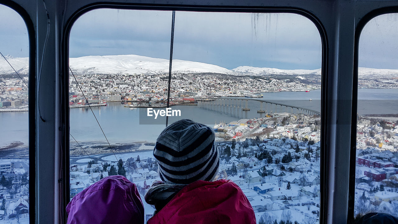 REAR VIEW OF WOMAN LOOKING THROUGH WINDOW AT WINTER