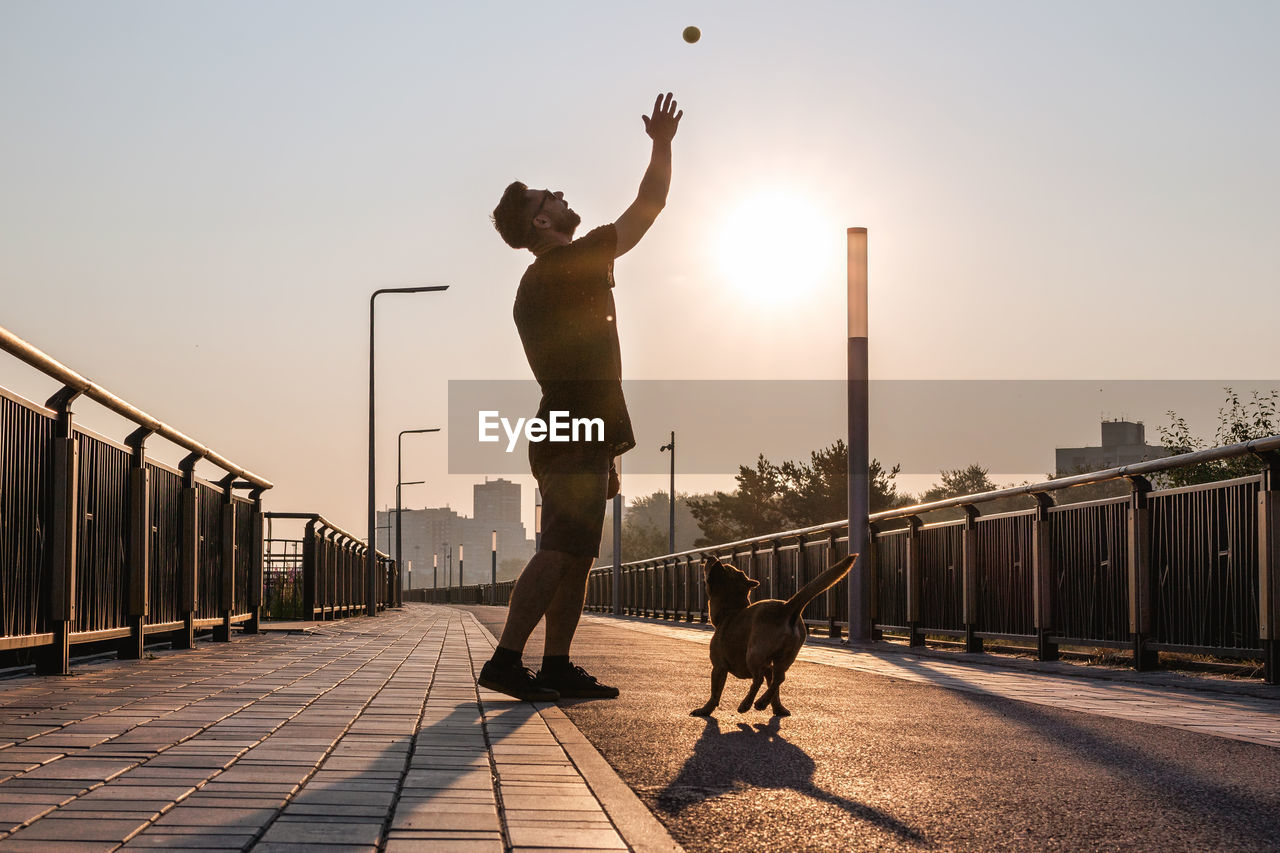 Young man is playing with his dog on empty street in the morning in backlight.