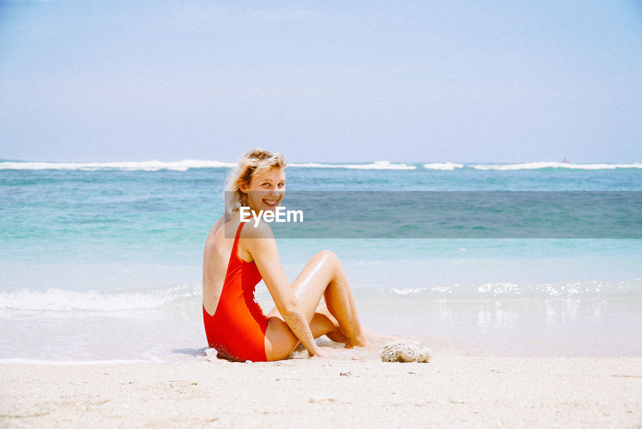 Portrait of smiling young woman sitting at beach against sky