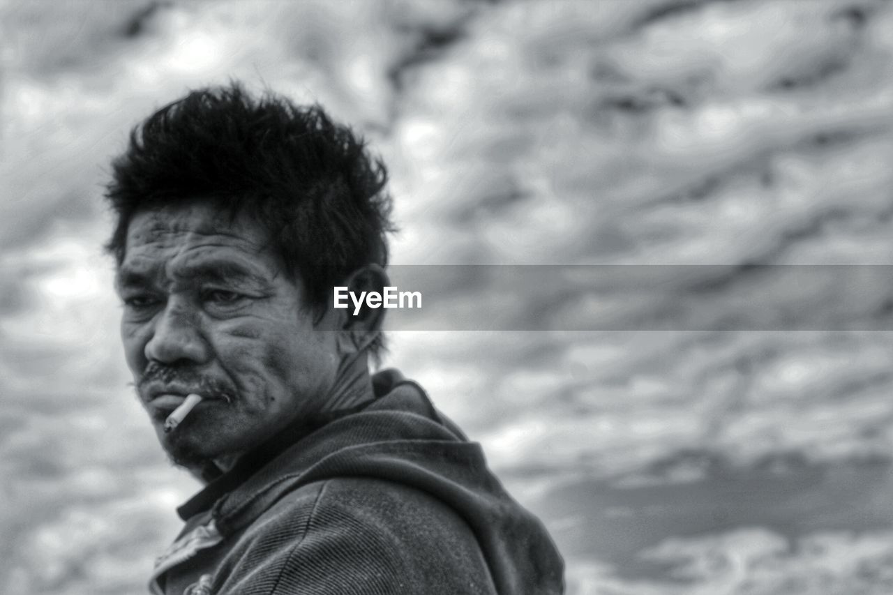 Side view of mature man smoking while standing against cloudy sky