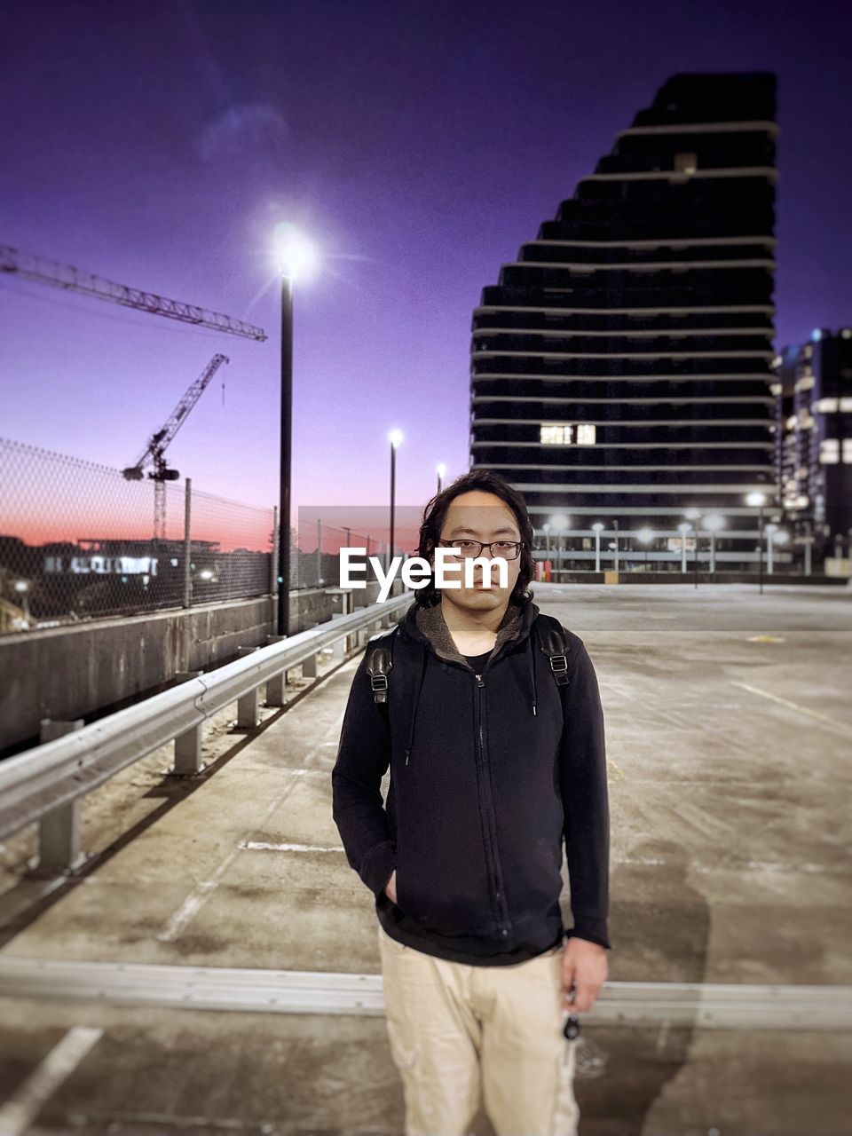 Portrait of young asian man standing on rooftop parking lot against lights and skyscrapers.