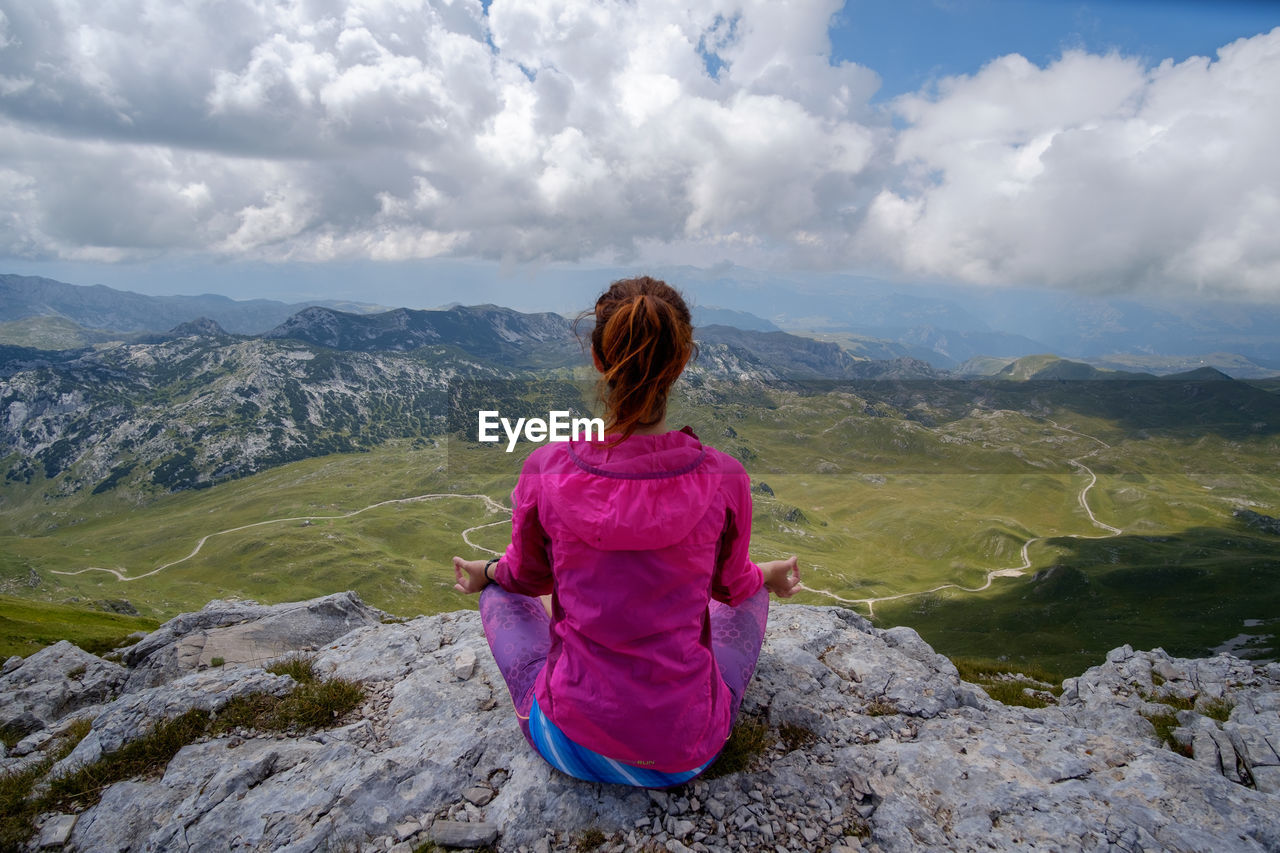 REAR VIEW OF WOMAN SITTING ON LANDSCAPE AGAINST MOUNTAIN