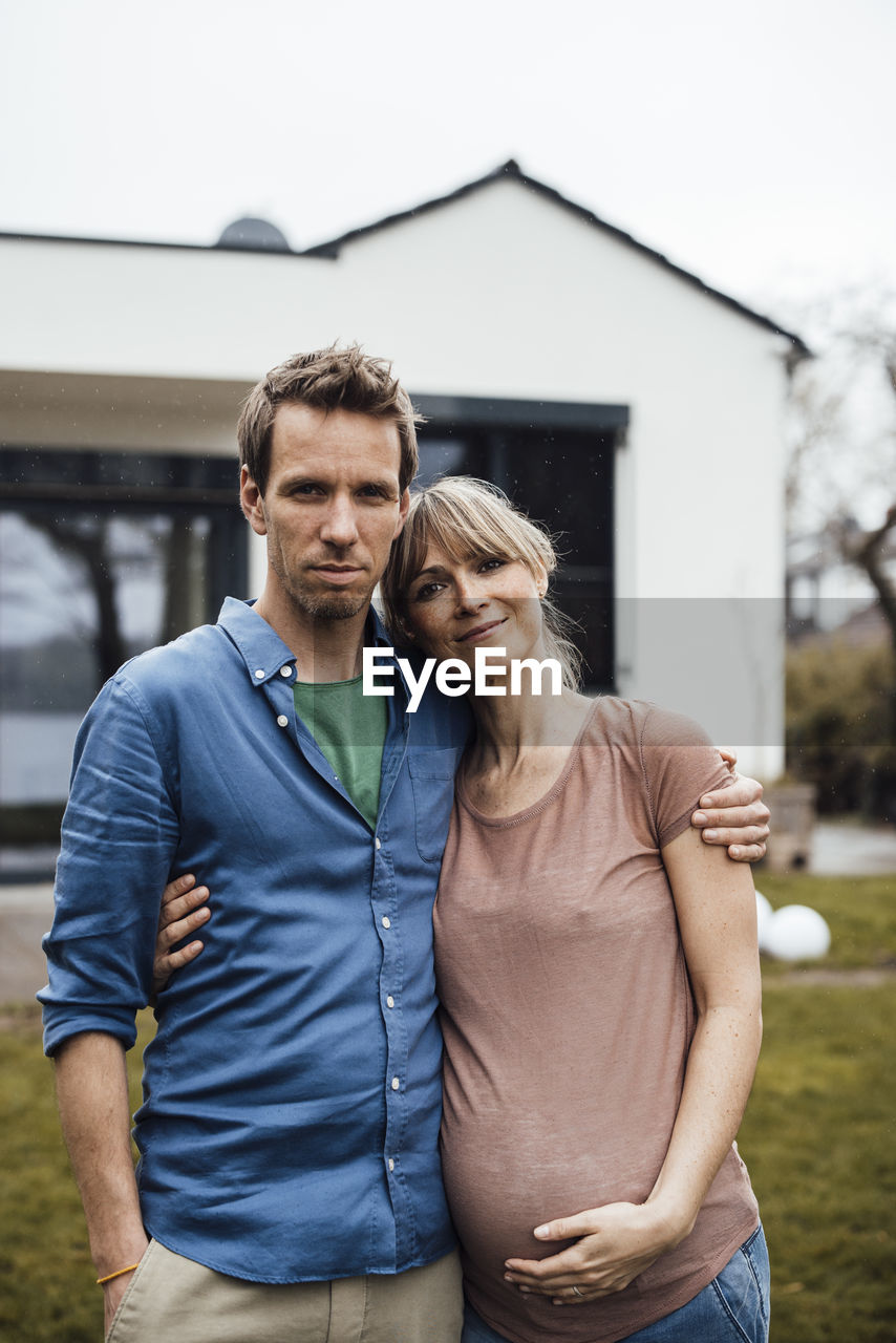 Expectant couple with arm around each other in front of house