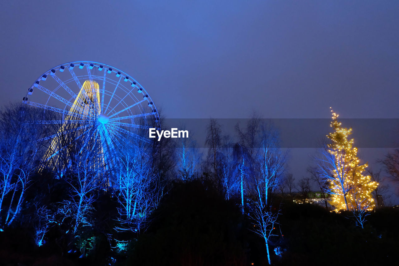 Low angle view of illuminated ferris wheel and bare trees against clear sky at dusk
