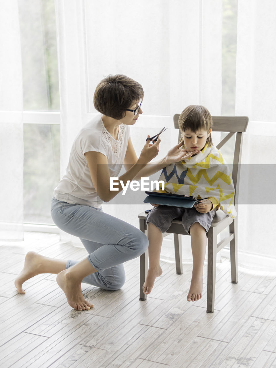 Mother cuts her son's hair by herself. little boy sits and holds digital tablet. new normal