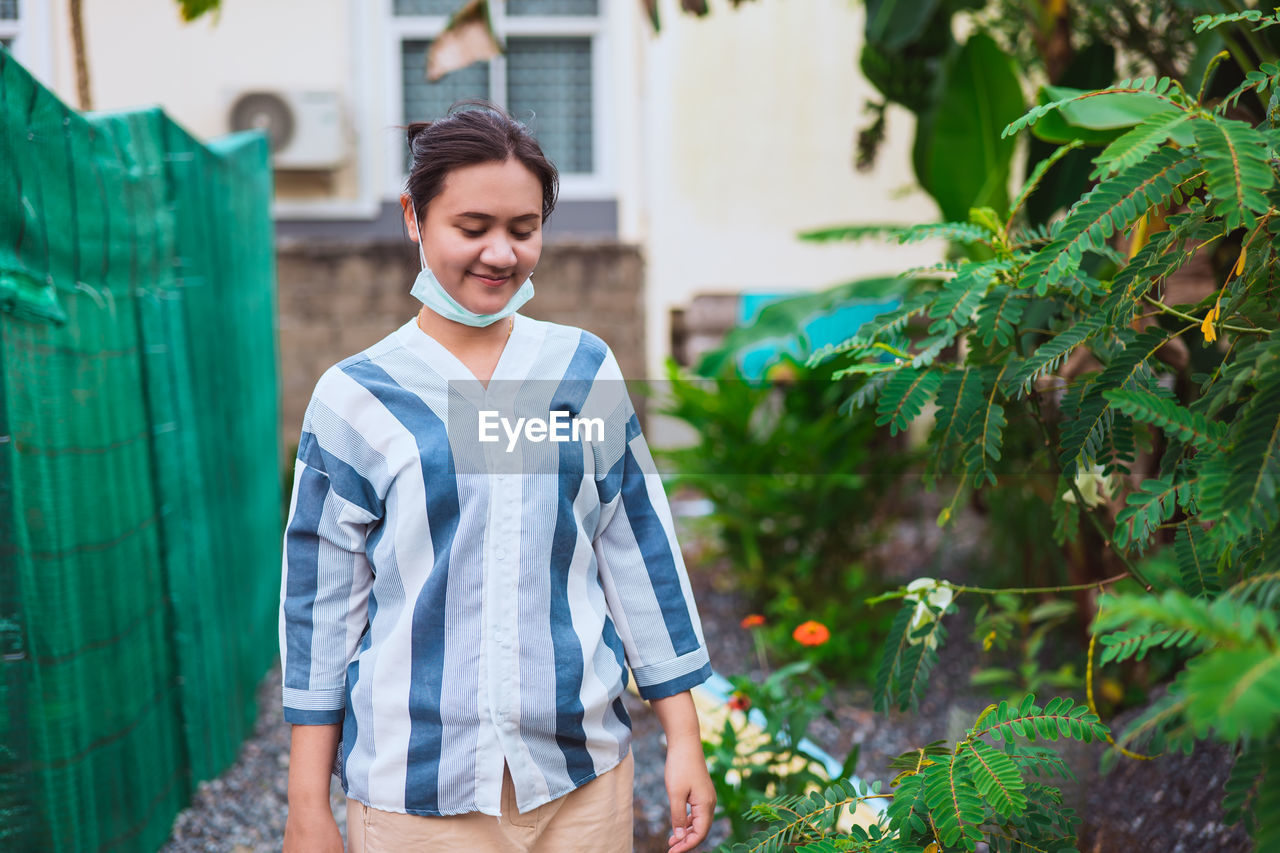 Asian woman walks through her own garden next to  house. there are green trees, green shading nets