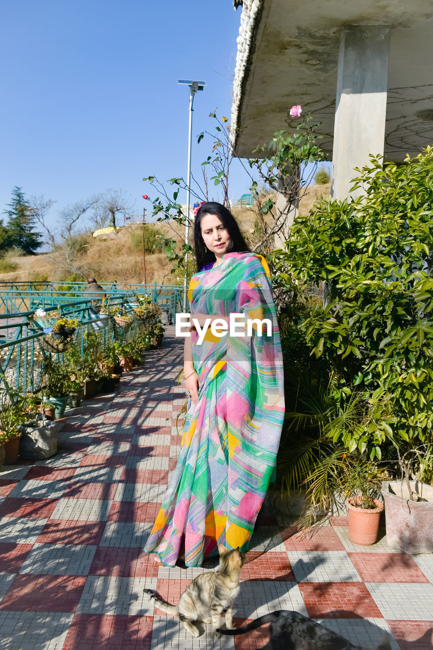 one person, adult, spring, full length, women, nature, architecture, young adult, plant, smiling, portrait, day, standing, clothing, dress, looking at camera, happiness, building exterior, vacation, flower, lifestyles, sunlight, fashion, built structure, outdoors, multi colored, sky, blue, leisure activity, yellow, green, traditional clothing, emotion, sunny, female, front view, building, casual clothing