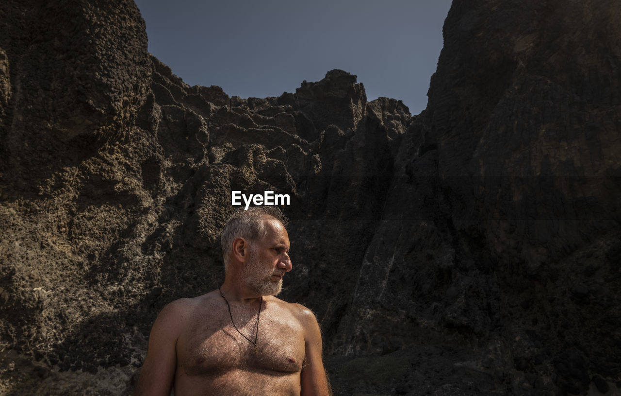 Portrait of shirtless man standing on beach against rock