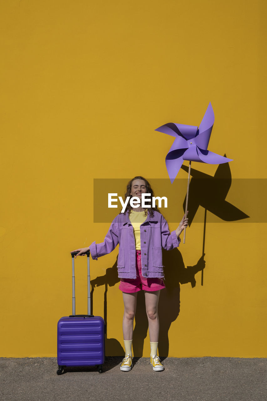 Smiling woman with purple luggage and pinwheel toy standing in front of yellow wall