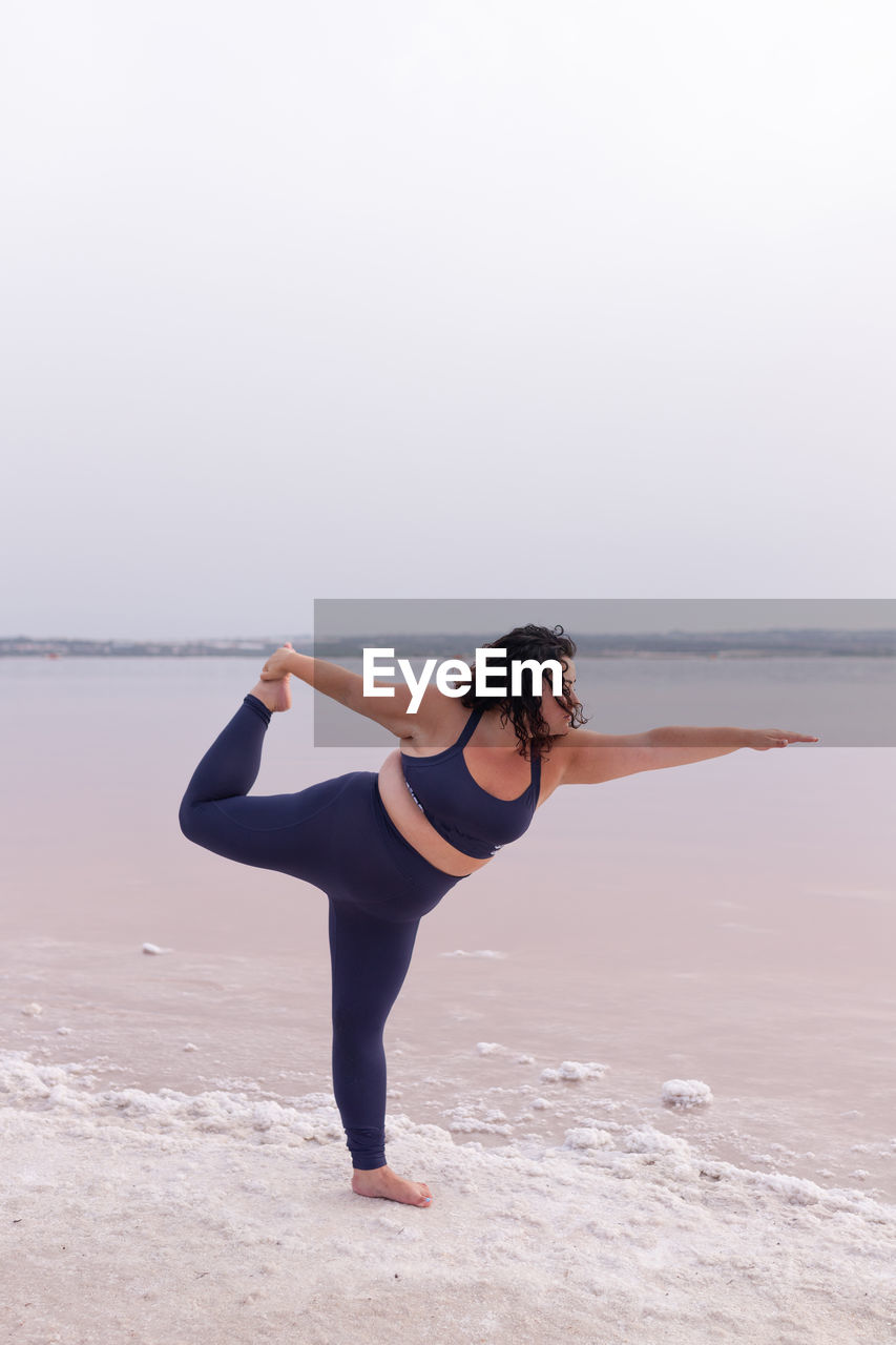 Side view of curvy female in sportswear balancing in natarajasana on shore of pink pond and practicing yoga in summer