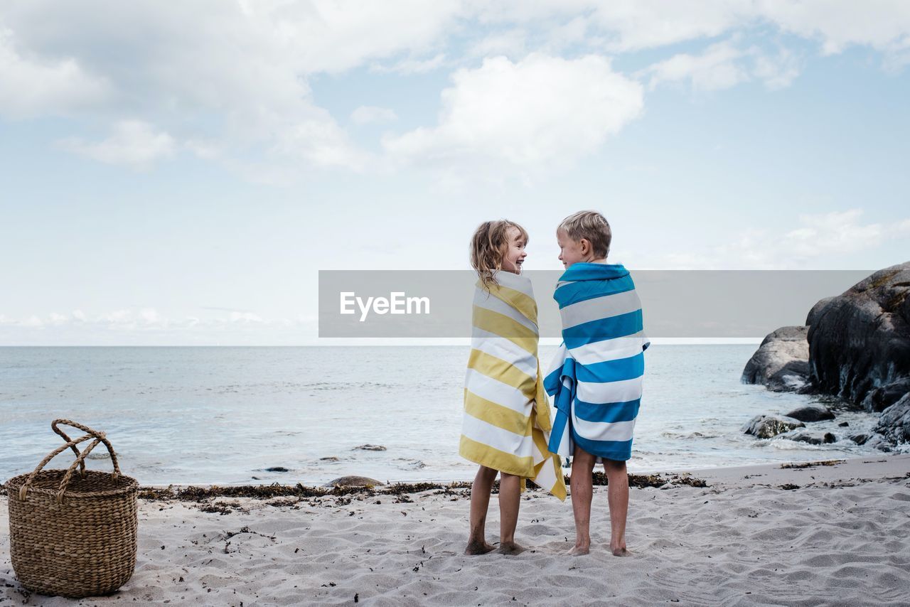 Brother and sister stood laughing at the beach wrapped in towels