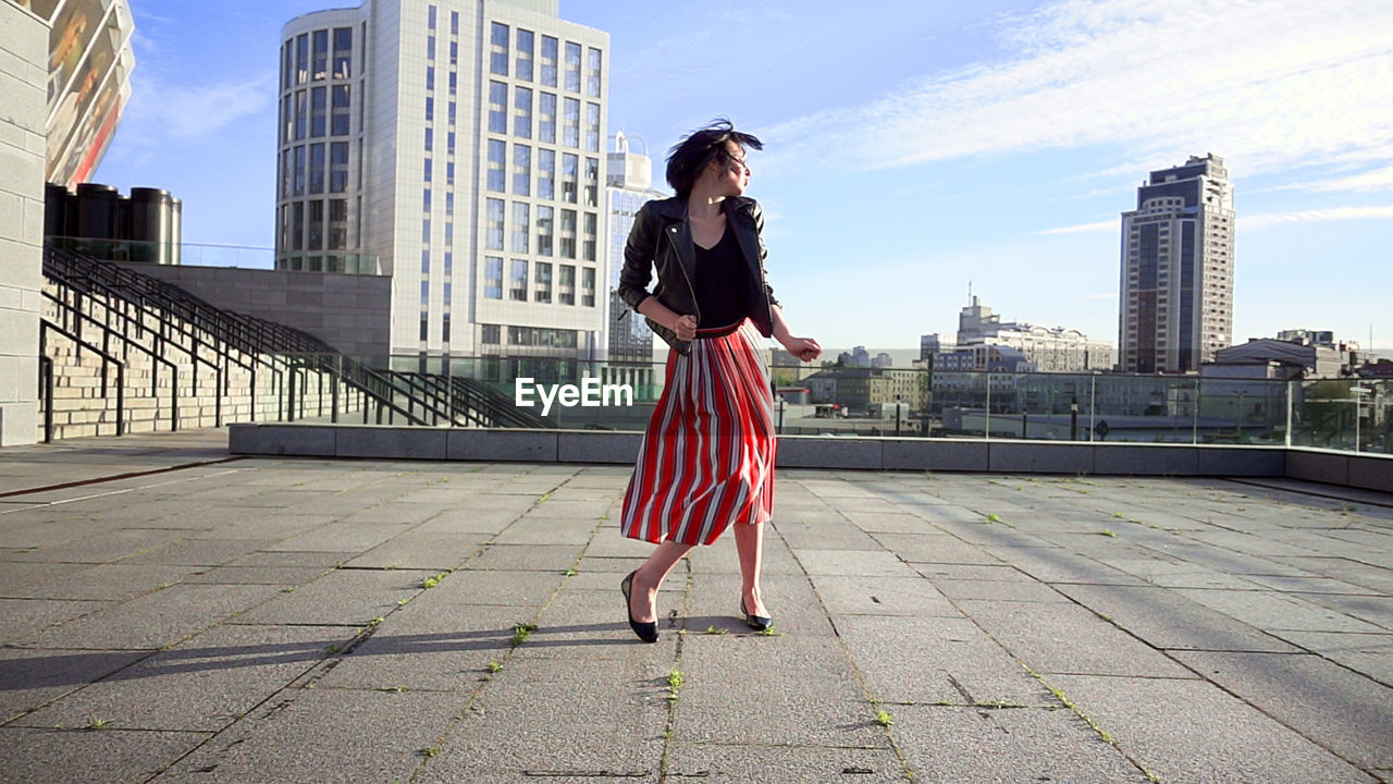 Happy woman wearing striped skirt and jacket on footpath in city