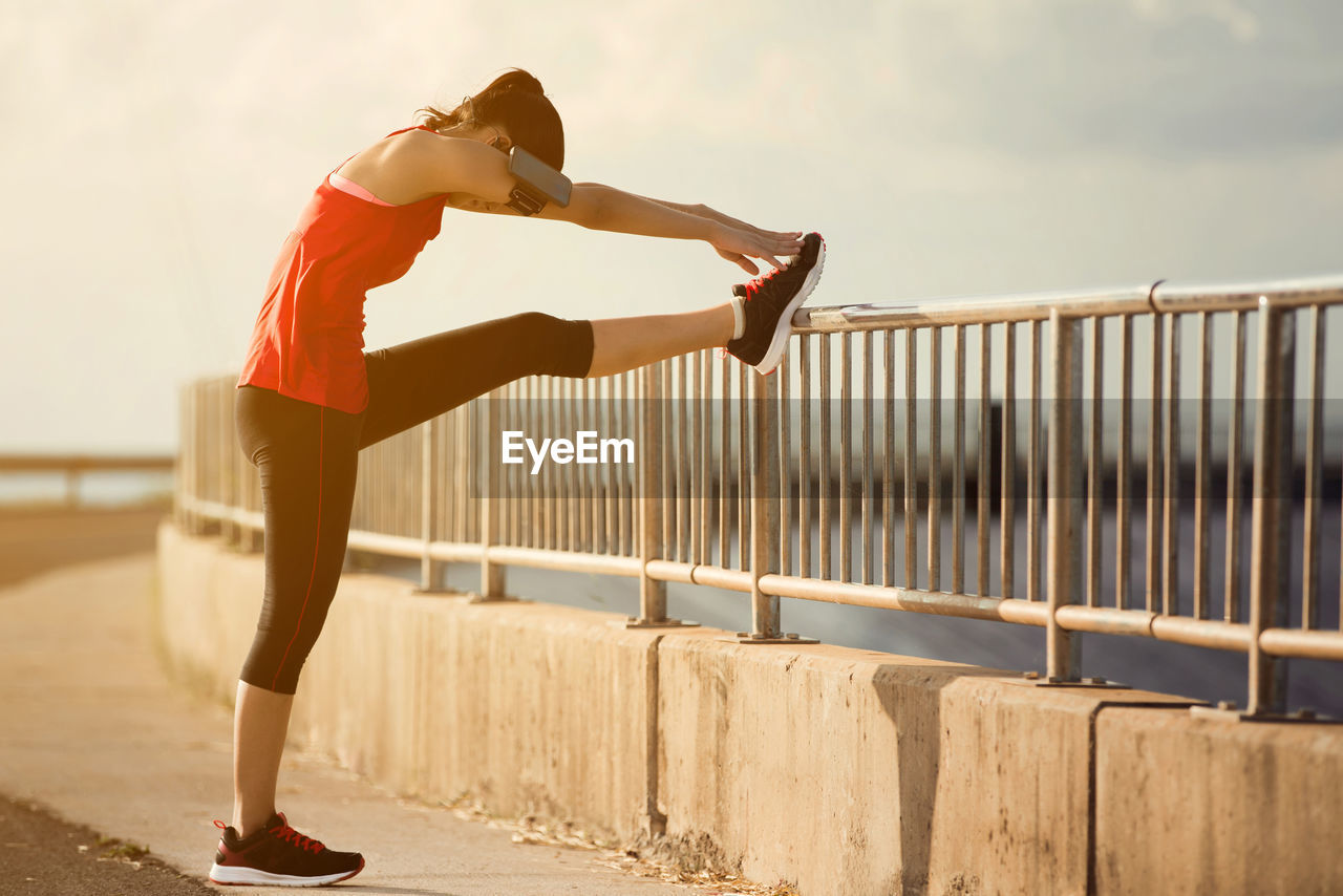 Side view of young woman exercising on railing against sky during sunset