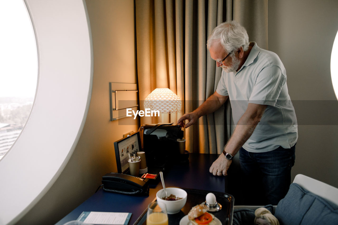 Senior man taking drink from espresso maker while standing by table in hotel room