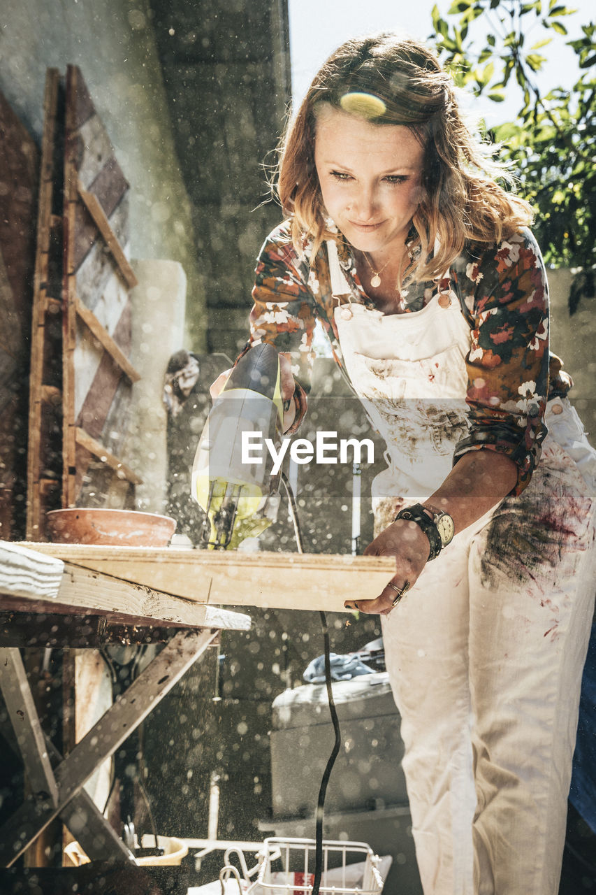 Female artist cutting wooden plank while working at backyard