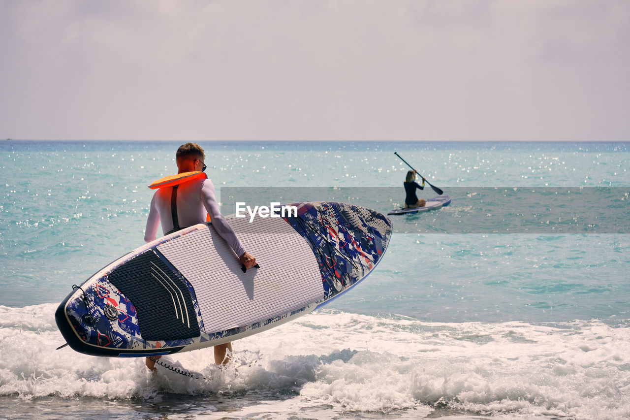 Young man dressed in swimsuit and lifevest carrying sup board towards sea