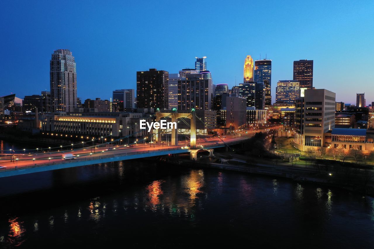 Illuminated buildings by river against clear sky at night